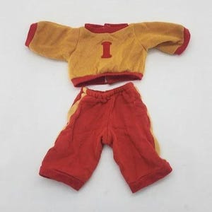 May include: A doll-sized outfit in gold and red. The top is gold with red trim and a large red "I" on the front. The matching shorts are red with gold stripes down the sides. The outfit is made of a soft fabric.