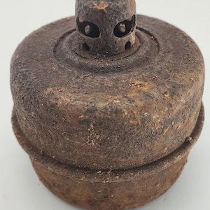 May include: A rusty, antique metal object with a cylindrical top featuring two eye-shaped holes and several smaller circular holes. The base is a wider, circular shape. The object has a weathered, aged appearance.