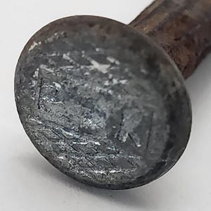 May include: Close-up of a weathered, gray metal nail with a rounded head. The head has a textured surface with visible wear and tear. The nail's shank is a darker, rusty brown color, suggesting age and exposure to the elements.