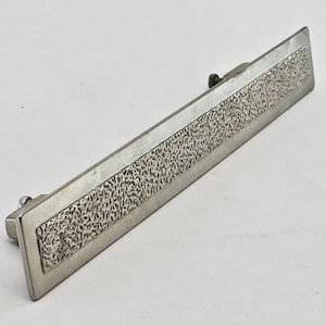 May include: A rectangular, silver-toned metal door pull with a textured center panel. The door pull has a simple, minimalist design, with a slightly raised, textured pattern in the center. The hardware is likely for a cabinet or drawer.