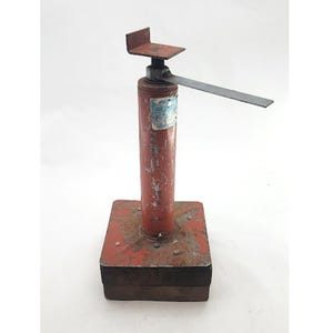 May include: A vintage red and metal jack stand with a rectangular base and a vertical, cylindrical body. The stand has a metal plate on top and a long, flat metal arm extending from the side. The paint is worn, showing signs of age and use.