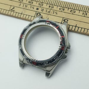 May include: A silver-toned watch bezel with a black and red rotating dial. The bezel has markings for minutes and seconds, with numbers in red and white. The watch bezel is a replacement part.