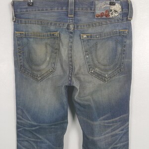 May include: Faded blue denim jeans with distressed details. Features a classic five-pocket design and a leather patch with a skull and rose graphic. The back pockets have a signature horseshoe-shaped stitching.