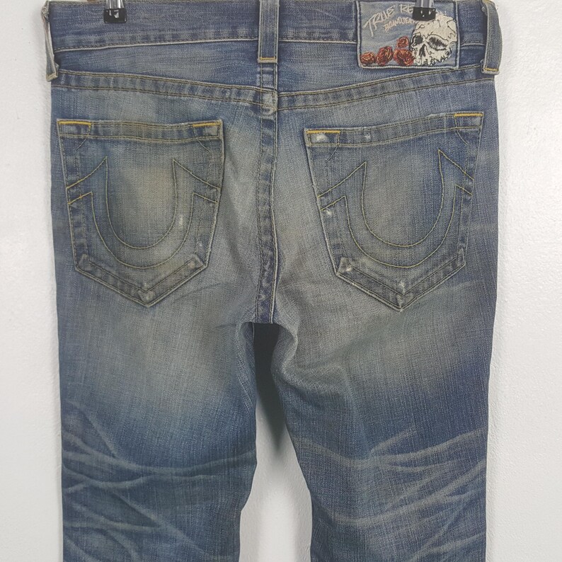 May include: Faded blue denim jeans with distressed details. Features a classic five-pocket design and a leather patch with a skull and rose graphic. The back pockets have a signature horseshoe-shaped stitching.