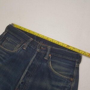 May include: Dark blue denim jeans featuring a metal button closure, front pockets, and copper-toned rivets. A yellow measuring tape is positioned along the waistband, indicating a width of approximately 15.75 inches across the top edge.