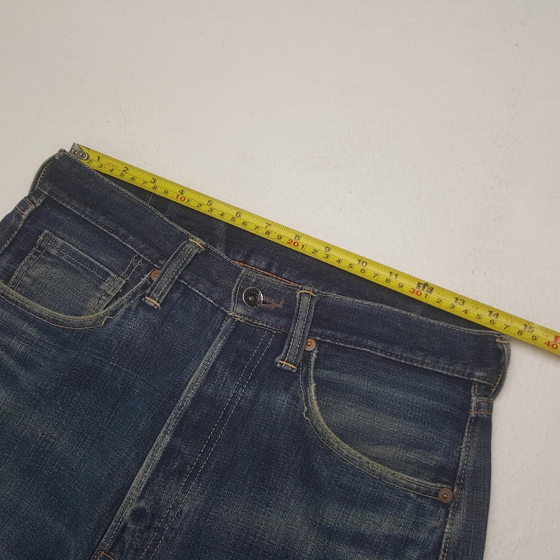 May include: Dark blue denim jeans featuring a metal button closure, front pockets, and copper-toned rivets. A yellow measuring tape is positioned along the waistband, indicating a width of approximately 15.75 inches across the top edge.