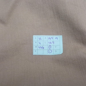 May include: A light brown fabric with a diagonal twill weave texture. A small light blue rectangular tag is attached, featuring a grid with handwritten text including C 22, A 19.5, N 4, L 26.5, W 446, and circled letters G and E.