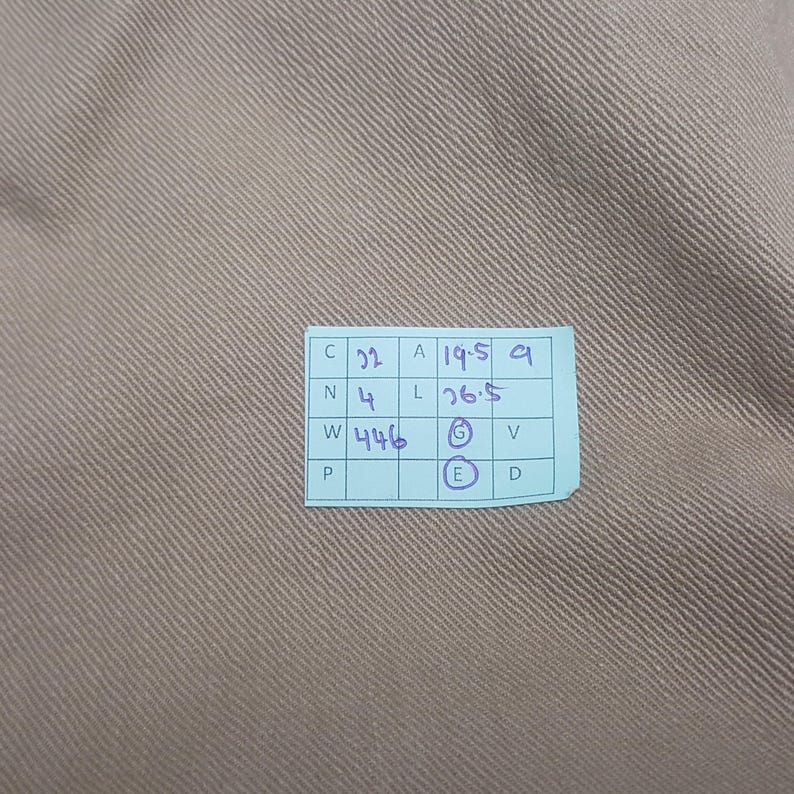 May include: A light brown fabric with a diagonal twill weave texture. A small light blue rectangular tag is attached, featuring a grid with handwritten text including C 22, A 19.5, N 4, L 26.5, W 446, and circled letters G and E.