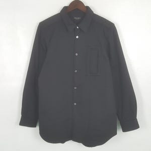Puede incluir: Camisa negra con botones, cuello y un bolsillo en el pecho. La camisa de manga larga está hecha de un material acolchado y cuelga de una percha de madera.