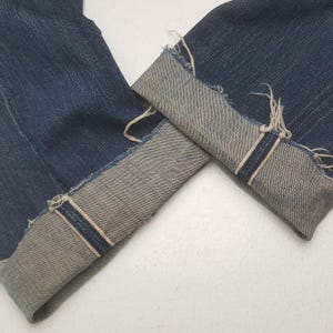 May include: Dark blue denim jeans featuring cuffed hems with a visible light-colored selvedge edge. The cuffs display a frayed, raw-edge finish along the fold, showcasing the contrast between the deep indigo exterior and the lighter interior fabric.