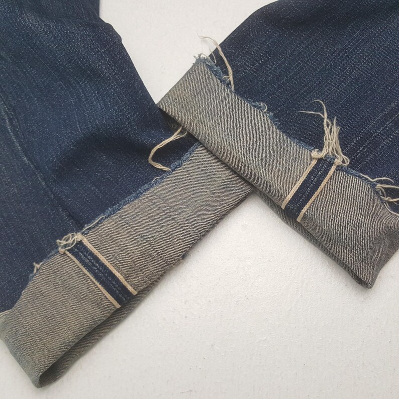 May include: Dark blue denim jeans featuring cuffed hems with a visible light-colored selvedge edge. The cuffs display a frayed, raw-edge finish along the fold, showcasing the contrast between the deep indigo exterior and the lighter interior fabric.