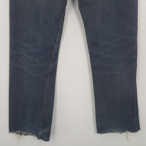 May include: A pair of dark blue denim jeans featuring a straight leg cut and a raw, frayed hem at the bottom of each leg. The fabric displays subtle whiskering and fading patterns across the upper thigh area, consistent with worn denim.