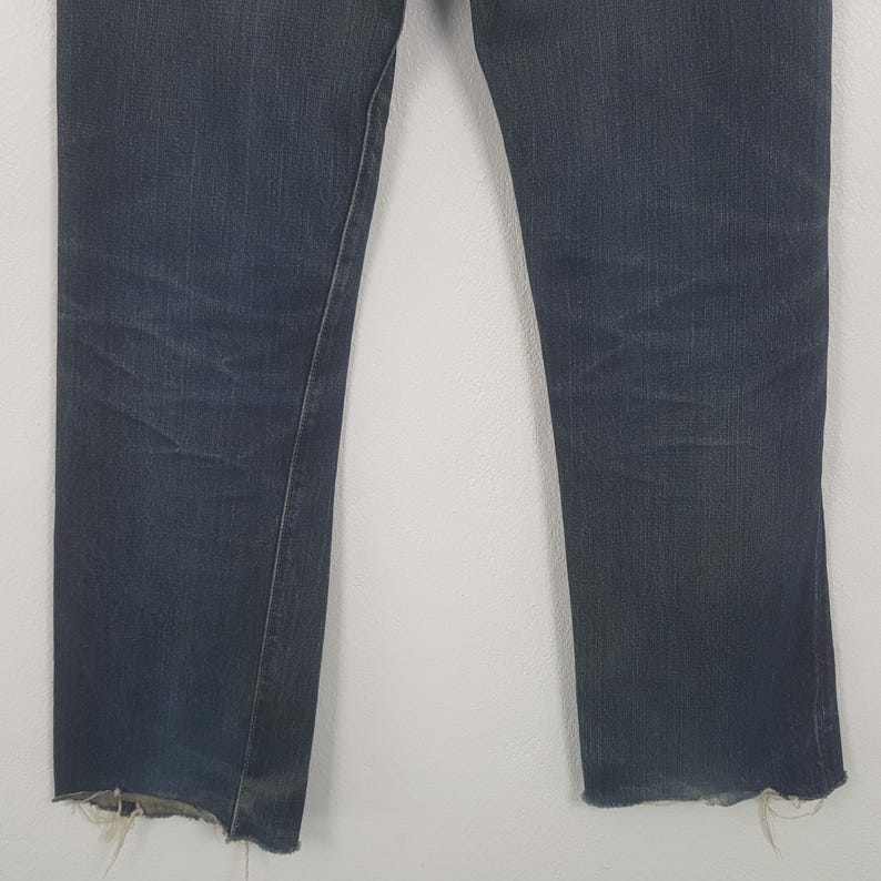 May include: A pair of dark blue denim jeans featuring a straight leg cut and a raw, frayed hem at the bottom of each leg. The fabric displays subtle whiskering and fading patterns across the upper thigh area, consistent with worn denim.