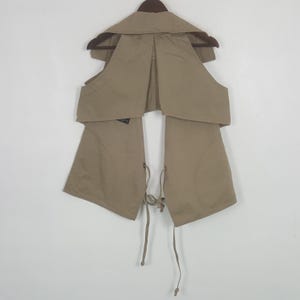 May include: A beige sleeveless vest featuring a wide collar, an open front design, and thin fabric ties at the waist. The garment is constructed from a solid-colored fabric and is displayed on a dark brown hanger against a plain white background.