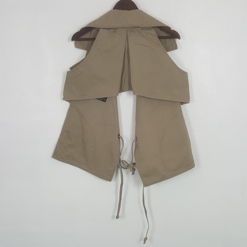 May include: A beige sleeveless vest featuring a wide collar, an open front design, and thin fabric ties at the waist. The garment is constructed from a solid-colored fabric and is displayed on a dark brown hanger against a plain white background.
