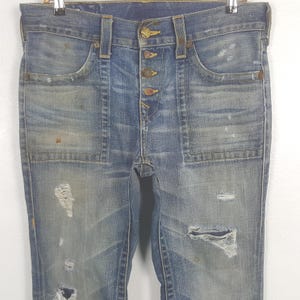 May include: Faded blue denim jeans with a button fly and distressed details. The jeans feature front pockets, back pockets, and a worn appearance with rips and patches. The jeans are hanging on a metal hanger.