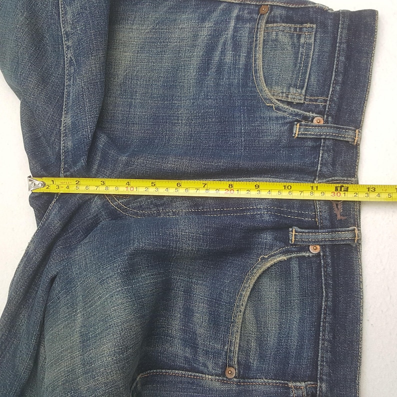 May include: A pair of blue denim jeans with a faded wash, featuring a classic five-pocket design, copper-toned rivets, and yellow stitching. A yellow measuring tape is placed across the waist, showing a width of approximately 13.5 inches.
