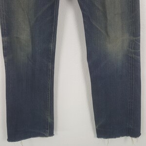 May include: A pair of dark blue denim jeans featuring a straight-leg cut with visible fading on the thighs and knees. The hem of each pant leg is finished with a raw, frayed edge, showing loose threads along the bottom of the denim fabric.