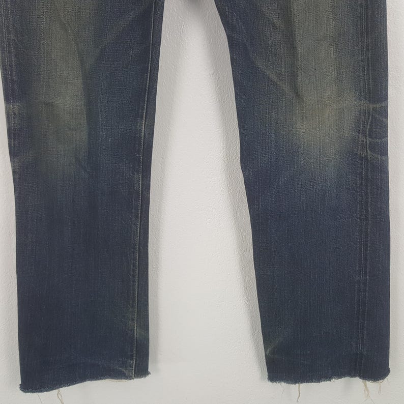 May include: A pair of dark blue denim jeans featuring a straight-leg cut with visible fading on the thighs and knees. The hem of each pant leg is finished with a raw, frayed edge, showing loose threads along the bottom of the denim fabric.