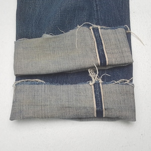 May include: Dark blue denim jeans featuring a cuffed hem with a visible selvedge edge. The fabric displays a classic twill weave texture, and the cuffs show frayed, raw edges along the bottom, highlighting the interior construction of the denim.