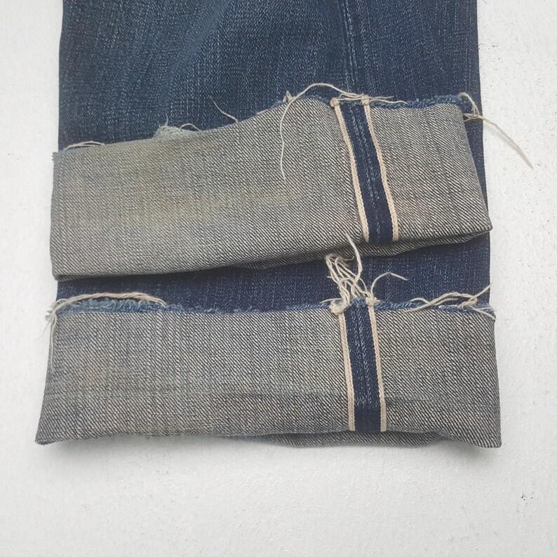 May include: Dark blue denim jeans featuring a cuffed hem with a visible selvedge edge. The fabric displays a classic twill weave texture, and the cuffs show frayed, raw edges along the bottom, highlighting the interior construction of the denim.