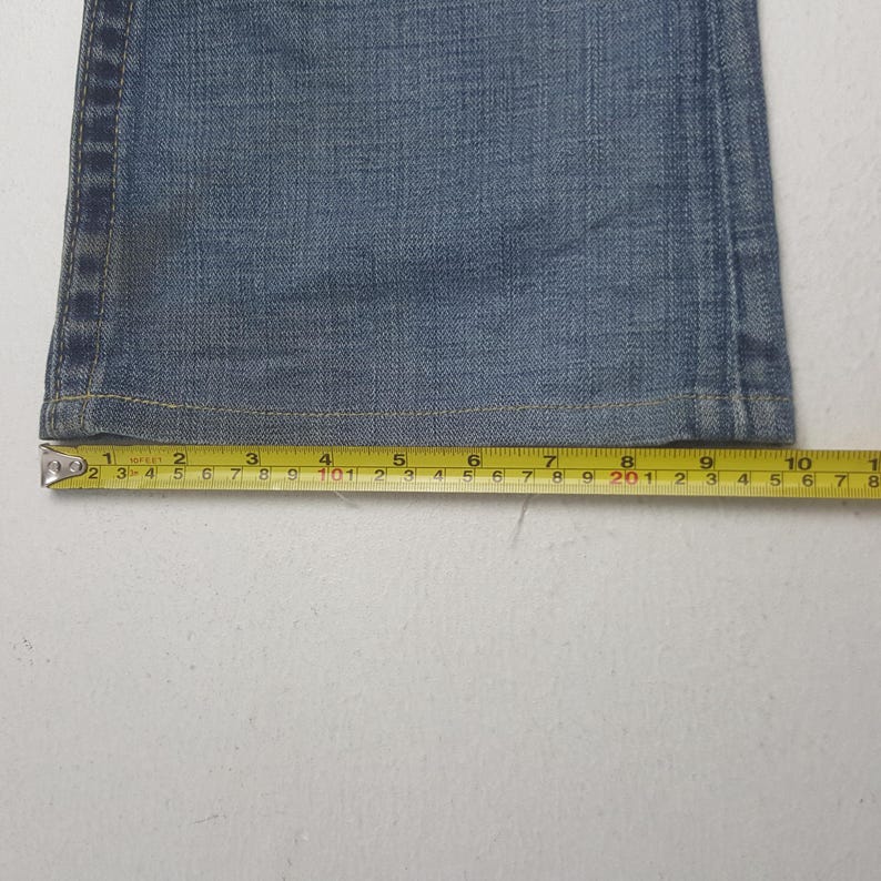 May include: Close-up of a section of blue denim jeans with a measuring tape. The tape shows measurements in inches, highlighting the fabric's texture and the jeans' construction details. The jeans are a light blue wash.