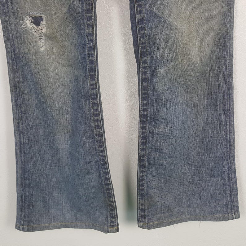 May include: Close-up of a pair of faded blue denim jeans. The jeans have a distressed look with a ripped section on the left leg. The jeans have a classic five-pocket design and a straight leg cut.