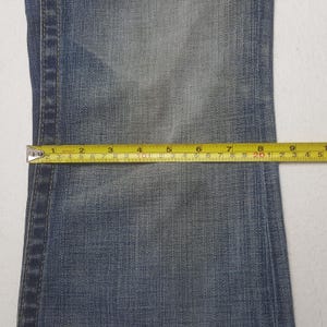 May include: Close-up of denim fabric with a measuring tape. The tape shows measurements in inches, highlighting the dimensions of the jeans. The fabric is a faded blue color.