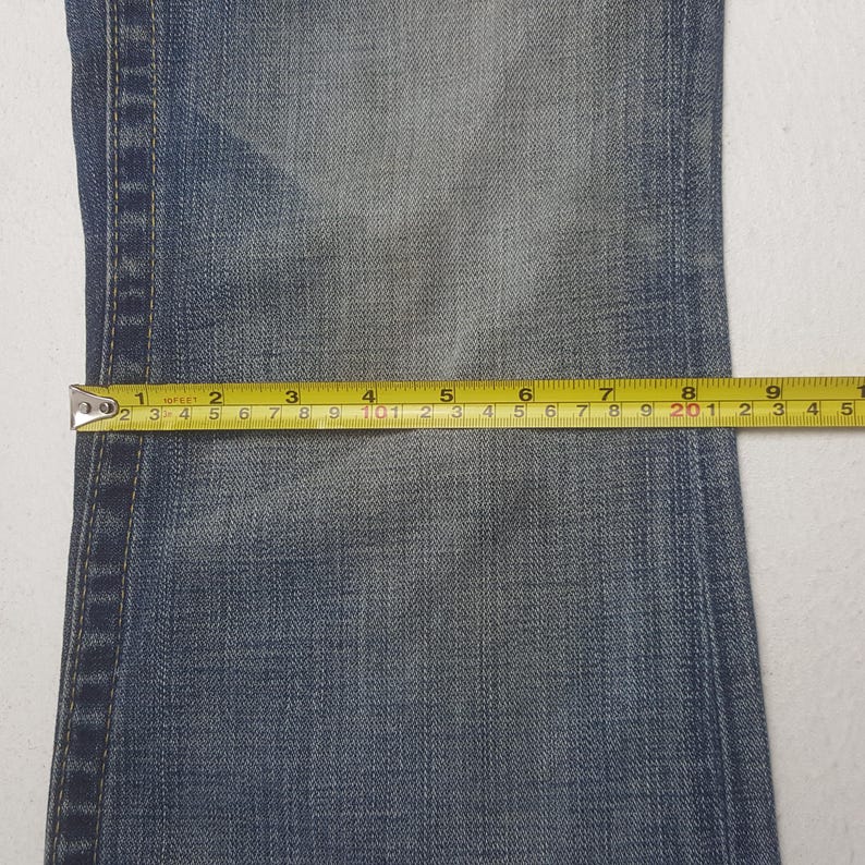 May include: Close-up of denim fabric with a measuring tape. The tape shows measurements in inches, highlighting the dimensions of the jeans. The fabric is a faded blue color.