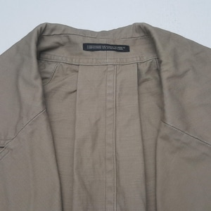 May include: A tan-colored jacket featuring a structured collar and a central vertical seam. A small black rectangular label is sewn into the inner neckline, containing the text: COMPOSITION SUR ETIQUETTE ANNEXE COMPOSITION SEE ATTACHED LABEL.