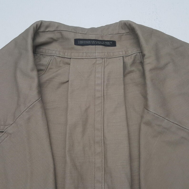 May include: A tan-colored jacket featuring a structured collar and a central vertical seam. A small black rectangular label is sewn into the inner neckline, containing the text: COMPOSITION SUR ETIQUETTE ANNEXE COMPOSITION SEE ATTACHED LABEL.