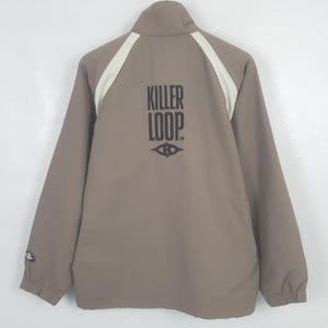 Op de afbeelding: Taupekleurige jas met witte schouderstrepen en een hoge kraag. De achterkant toont de zwarte tekst "KILLER LOOP" en een gestileerd ooglogo. De jas heeft lange mouwen en een ritssluiting.