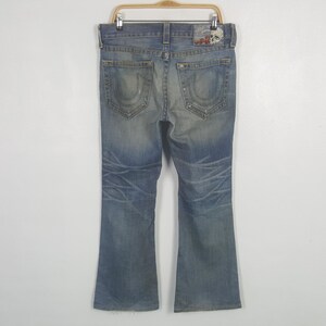 May include: Faded blue denim jeans with a bootcut leg. The jeans have a distressed look with fading and whiskering details. Features include back pocket stitching and a leather patch with a skull design.