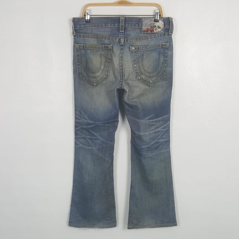 May include: Faded blue denim jeans with a bootcut leg. The jeans have a distressed look with fading and whiskering details. Features include back pocket stitching and a leather patch with a skull design.