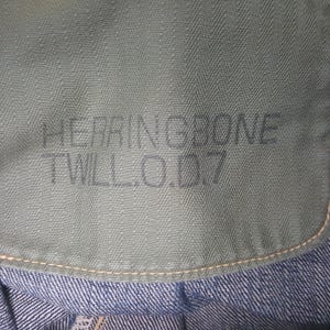 May include: A sage green fabric panel featuring a distinct herringbone weave pattern. The text HERRINGBONE TWILL.O.D7 is printed in a dark, sans-serif font across the center. The fabric is stitched with light-colored thread along the bottom edge.