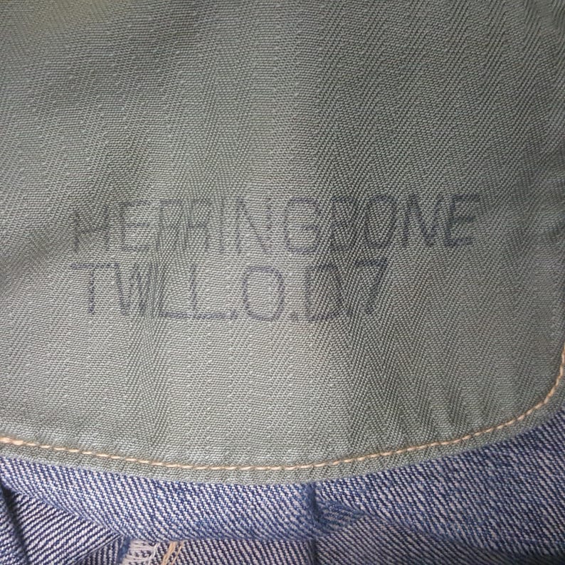 May include: A sage green fabric panel featuring a distinct herringbone weave pattern. The text HERRINGBONE TWILL.O.D7 is printed in a dark, sans-serif font across the center. The fabric is stitched with light-colored thread along the bottom edge.