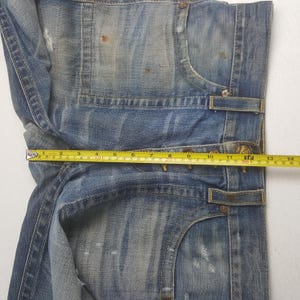 May include: Close-up of faded blue denim jeans with distressed details. A yellow measuring tape shows a length of approximately 13 inches. The jeans have visible wear and tear, with a classic five-pocket design.
