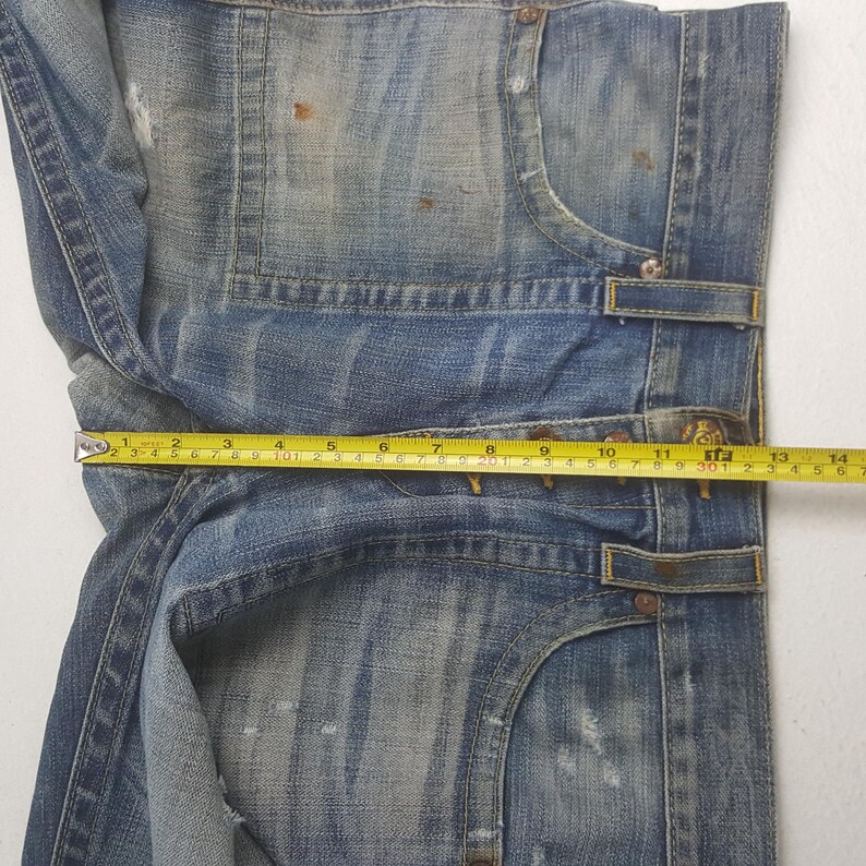 May include: Close-up of faded blue denim jeans with distressed details. A yellow measuring tape shows a length of approximately 13 inches. The jeans have visible wear and tear, with a classic five-pocket design.