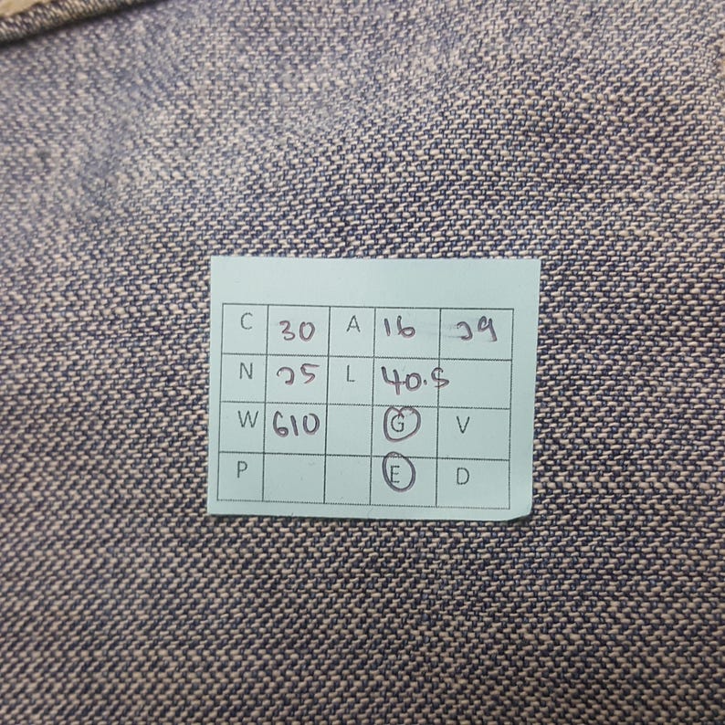 May include: A close-up of a blue and gray textured fabric. A small, rectangular, light green tag is affixed to the fabric. The tag contains black text and numbers, including "C 30", "A 16", "29", "N 25", "L 40.5", and "W 610".