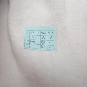 以下が含まれることがあります： 白い布のクローズアップで、青い付箋が付いています。付箋には、黒インクで手書きされた数字と文字が含まれており、「C 27」、「A 24」、および「L 26」が含まれています。生地は質感のある外観をしています。