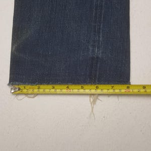 May include: A dark blue denim pant leg with a raw, frayed hem. A yellow measuring tape is positioned horizontally across the bottom edge, showing a width of approximately 9.5 inches from the left side to the right edge of the fabric.