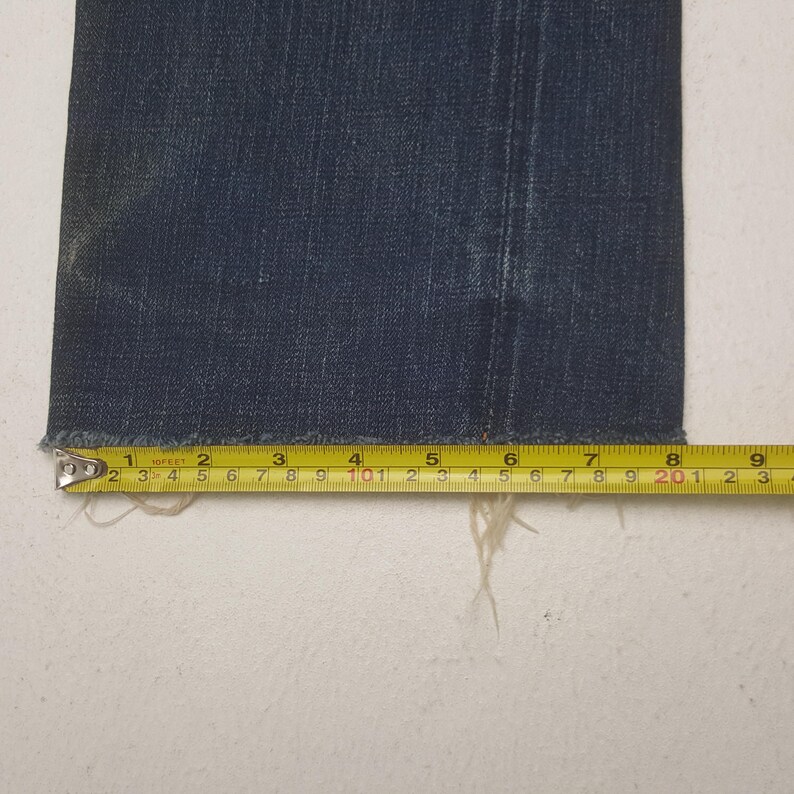 May include: A dark blue denim pant leg with a raw, frayed hem. A yellow measuring tape is positioned horizontally across the bottom edge, showing a width of approximately 9.5 inches from the left side to the right edge of the fabric.