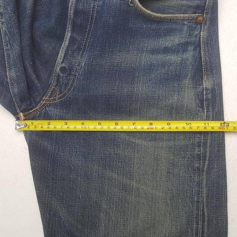 May include: A pair of dark blue denim jeans featuring a faded wash and visible stitching. A yellow measuring tape is placed across the fabric, indicating a width measurement of approximately 11.5 inches from the side seam to the crotch area.