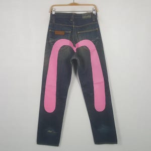 Puede incluir: Vaqueros de mezclilla azul oscuro con un gráfico grande y llamativo de una gaviota pintada en rosa sobre los bolsillos traseros. Los pantalones tienen una construcción clásica de cinco bolsillos, un parche de cuero marrón y un acabado desgastado.