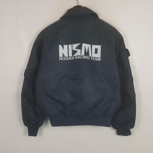 Puede incluir: Una chaqueta tipo bomber negra con cuello alto y puños acanalados. La parte posterior muestra el texto blanco NISMO y NISSAN RACING TEAM impreso en una fuente negrita sin remates. La chaqueta está hecha de un material sintético liso y sólido.