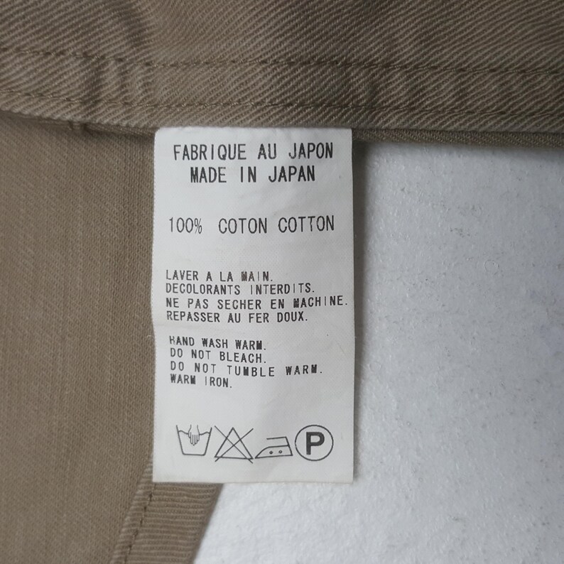 May include: A white rectangular garment care label sewn onto a tan fabric. The label features black text stating FABRIQUE AU JAPON, MADE IN JAPAN, 100% COTON COTTON, and care instructions including HAND WASH WARM, DO NOT BLEACH, DO NOT TUMBLE WARM, WARM IRON.