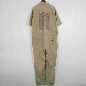 May include: A beige short-sleeved jumpsuit with a button-down collar and pockets. The back of the jumpsuit has a dark graphic design. The garment is suspended from a wooden hanger. The Dickies logo is visible on the back pocket.