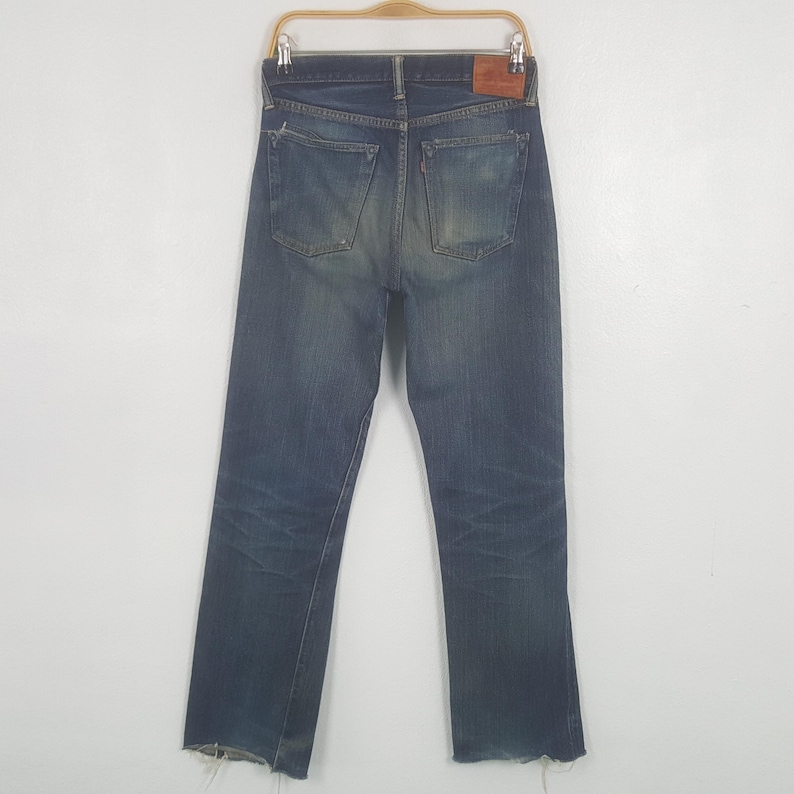 May include: A pair of blue denim jeans featuring a faded wash, two back pockets with reinforced stitching, a brown leather patch on the waistband, and a small red tab on the right pocket. The hem of the pant legs shows a frayed, raw edge finish.