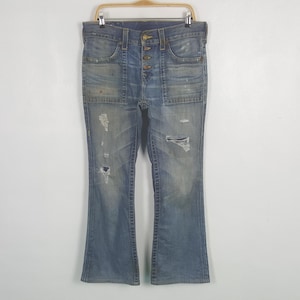 May include: Light blue denim jeans with a button fly and distressed details. The jeans have a flared leg and multiple pockets. The jeans are hanging on a wooden hanger against a white background.