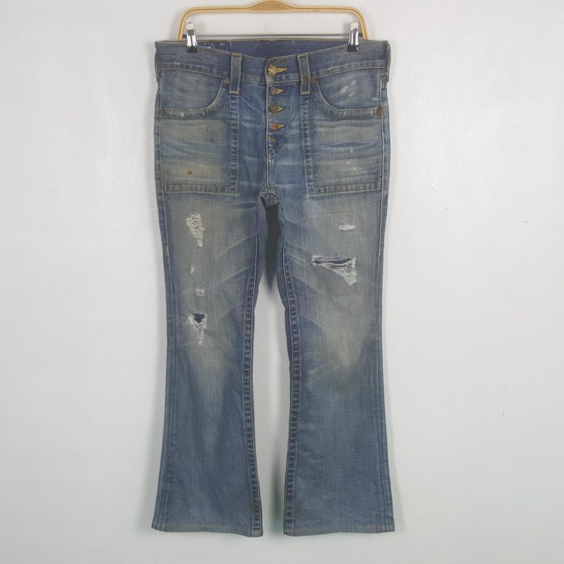 May include: Light blue denim jeans with a button fly and distressed details. The jeans have a flared leg and multiple pockets. The jeans are hanging on a wooden hanger against a white background.